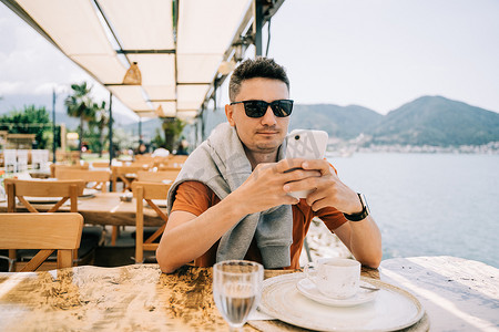 戴着墨镜的年轻男性在街道海边咖啡馆餐厅咖啡店里看着他的智能手机小工具，背景是风景秀丽的山脉。