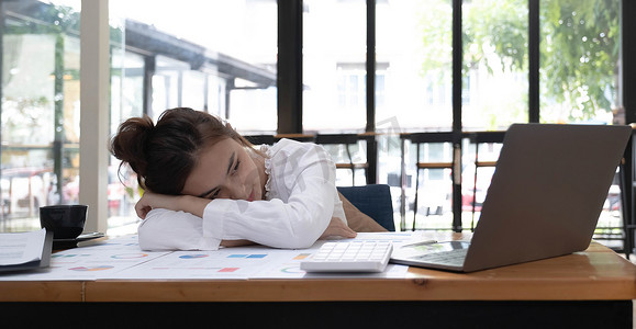 疲惫或疲惫的年轻女商人在办公室笔记本电脑前的办公桌上小睡