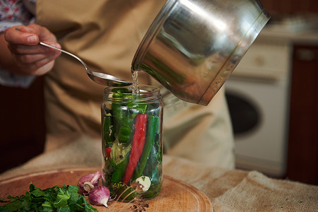 在家罐装时令蔬菜时，将沸腾的盐水或泡菜倒在玻璃罐中的辣椒上的特写