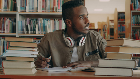 戴着大耳机的积极的非洲裔美国年轻帅哥坐在桌边，手里拿着书，透过窗户，微笑着，在他的文案上写字