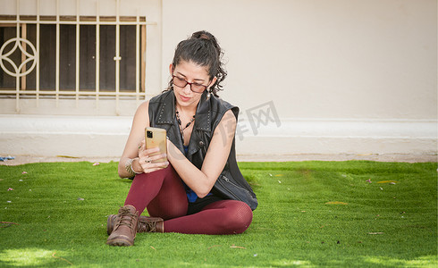 坐在草地上检查手机的女孩，坐在手机上的城市女孩，带手机的女性的生活方式