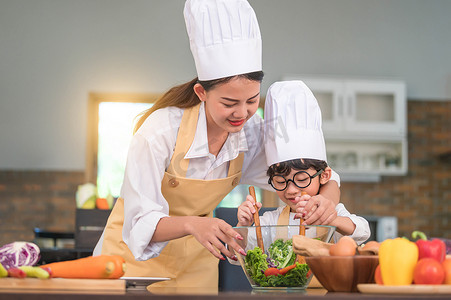 快乐美丽的亚洲女人和戴着眼镜的可爱小男孩准备在家里的厨房里做饭。