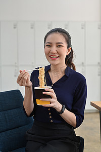 微笑的亚洲女人拿着塑料叉子在办公室吃方便面。
