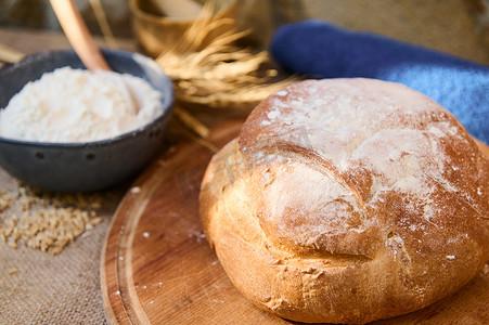 一条新鲜出炉的自制全麦酵母面包，放在木板上，碗里放着面粉和小麦小穗。