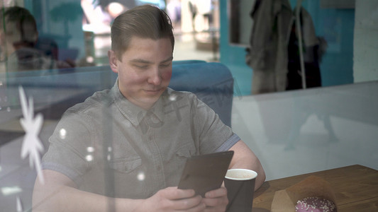 咖啡馆摄影照片_一个年轻人坐在咖啡馆的玻璃杯后面，手里拿着一台平板电脑。