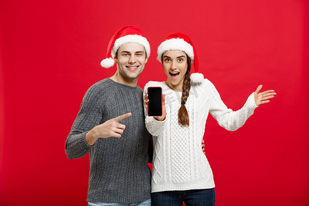 圣诞毛衣摄影照片_圣诞节概念 — 穿着圣诞毛衣的快乐年轻夫妇用手机指着手指