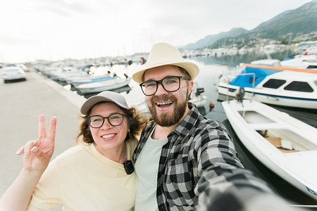 一对年轻夫妇在夏季港口码头的户外码头上摆着小船和游艇，一边用智能手机近距离拍照，一边笑着自拍
