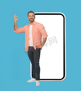 手势 OK 站在大智能手机与白色屏幕英俊的中年灰色头发的男人微笑着穿着桃色衬衫隔离在蓝色背景。