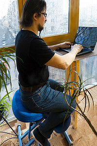 一名男子坐在阳台上的符合人体工程学的矫形跪椅上使用笔记本电脑工作。