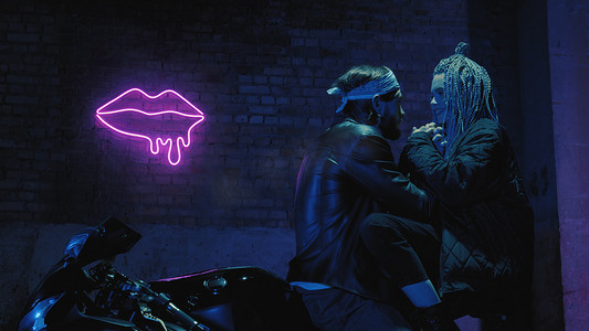 骑摩托的男人摄影照片_一个恋爱中的女孩和一个男人坐在超级运动摩托车上，在霓虹灯的背景下调情和拥抱