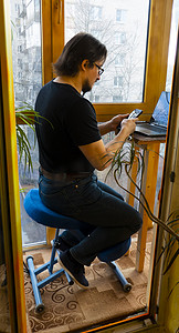 一名男子坐在阳台上的符合人体工程学的矫形跪椅上使用笔记本电脑工作。