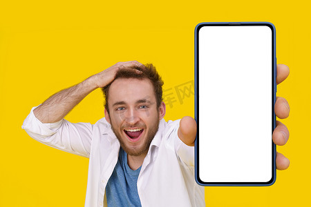 巨大手机摄影照片_英俊兴奋的年轻人情绪激动地微笑着摸着他的头，展示着带有白色屏幕的巨大智能手机。