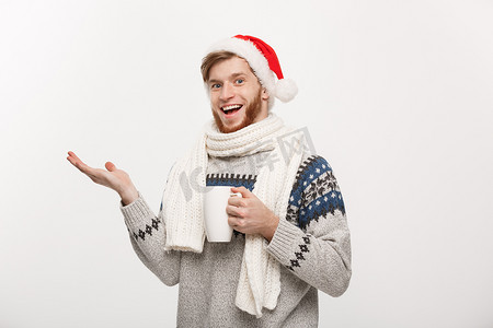 圣诞节概念 — 穿着毛衣和圣诞老人​​帽子的年轻胡子男子拿着一个热咖啡杯，手指向一侧，与白色隔离，有复制空间。