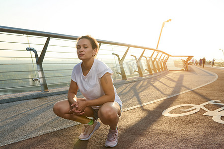 身着白色 T 恤和蓝色牛仔短裤的迷人中年西班牙裔女性的肖像蹲在跑步机上，看着相机，同时享受日出时的清晨散步