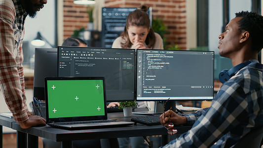 程序员摘下眼镜与办公桌前的同事交谈，笔记本电脑配有绿屏色度键模型
