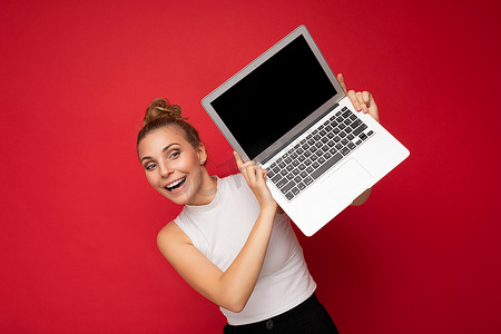 照片中，美丽、迷人、积极、微笑的年轻金发女子被隔离在红色背景墙上，空白的文字空间，穿着休闲白色上衣，手持带有黑色空白显示屏的笔记本电脑用于模型