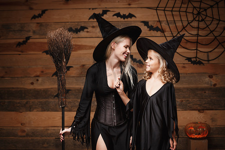 女巫蝙蝠摄影照片_万圣节概念-欢快的母亲和她的女儿穿着女巫服装庆祝万圣节，在木制工作室背景下与蝙蝠和蜘蛛网上的弯曲南瓜合影。