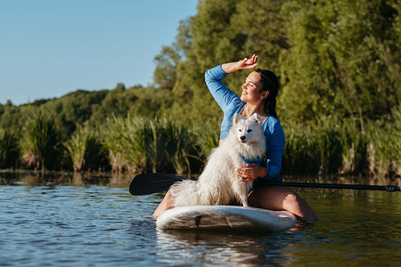 清晨，湖上快乐的年轻女子和她的狗雪白的日本斯皮茨坐在 Sup 板上，享受日出