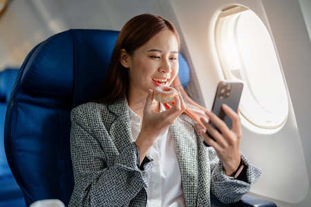 一位成功的亚洲女商人或女性企业家在飞机上穿着正式西装的肖像坐在商务舱座位上，在飞行期间吃饭和使用智能手机