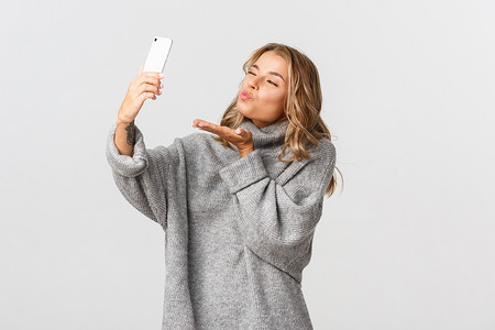 身穿灰色毛衣的漂亮年轻女子手持智能手机、自拍、在视频通话期间向手机摄像头发送飞吻、站在白色背景上的肖像