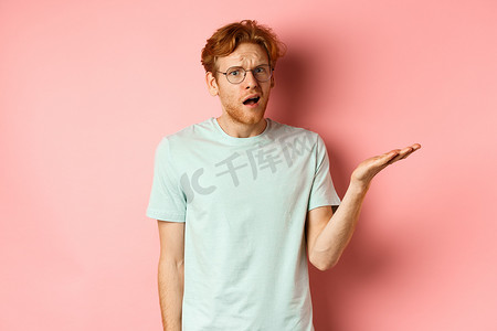 戴眼镜的英俊红发男子的肖像看起来很困惑，张开嘴，盯着相机，什么也听不懂，站在粉红色的背景上