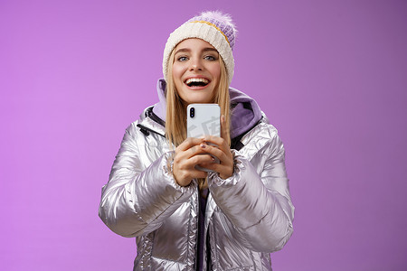 有趣兴奋的迷人金发女友举着智能手机录制视频男友踏滑雪板第一次捕捉记忆手机相机快乐地站在紫色背景
