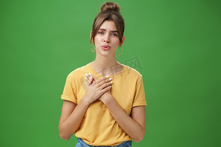 感恩摄影照片_身穿黄色 T 恤的迷人年轻女性以感恩和感激的姿态将手掌压在胸前，折叠嘴唇融化了心，看到绿色背景中的可爱事物