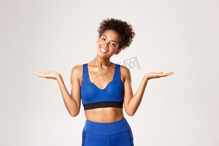 穿着蓝色运动服、微笑着的非洲裔美国健身女孩的腰部向上拍摄，侧身举起双手，拿着东西或示威，站在白色背景下