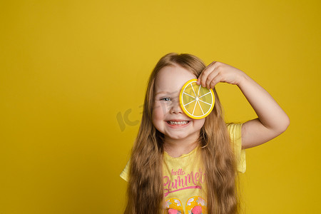 小女孩柠檬摄影照片_微笑的小女孩在眼睛附近拿着一片柠檬，看着相机中特写的肖像