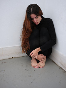 房间的角落摄影照片_一个害怕的年轻女人坐在房间的角落里，穿着黑色的衣服，赤脚往下看。