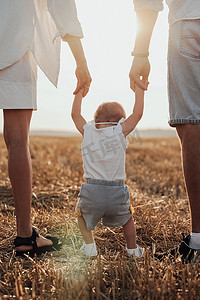 父母平安摄影照片_父母帮助孩子迈出第一步的特写后视图，年轻家庭在日落时在田野上行走