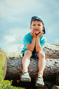 坐在树上的小孩摄影照片_一个戴着蓝色棒球帽和牛仔短裤的小孩坐在木头上，开心地笑着