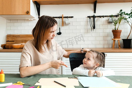 可爱生气的学龄前小女孩坐在客厅的桌子旁和妈妈一起做创造性的家庭作业并争论。