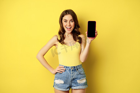 app展示界面摄影照片_时尚的微笑女性展示手机屏幕、智能手机上的应用程序界面、站在背心和黄色背景下的短裤