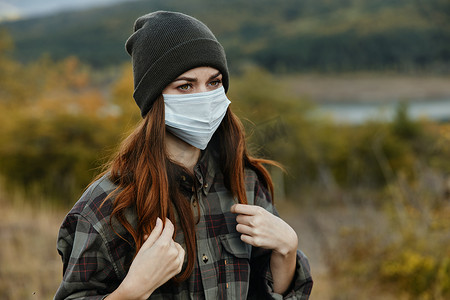 美丽的女人脸上戴着医用面具，在森林里的户外戴着温暖的帽子