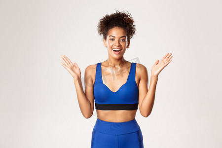 穿着蓝色运动服的惊讶而快乐、健康的非洲裔美国健身女孩的腰部，举起双手，高兴地微笑着，站在白色背景下