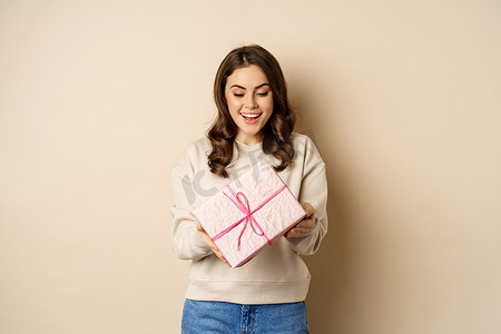 兴奋的漂亮女孩，带着粉色包裹的礼盒，收到礼物，站在米色背景上