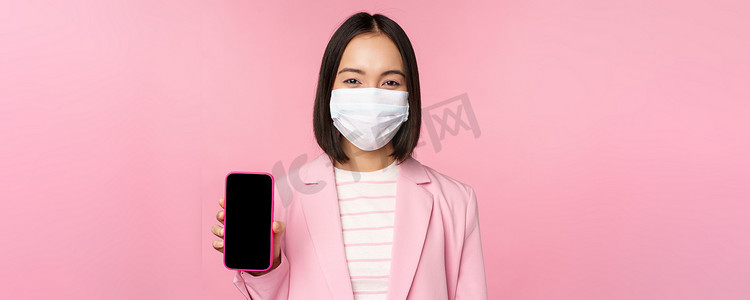 身着医用面罩、商务套装、展示智能手机屏幕、站在粉红色背景上的微笑韩国女售货员的画像