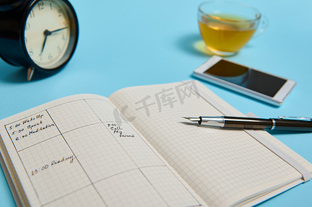 工作闹钟摄影照片_蓝色表面上有一个开放式日程表、智能手机、墨水笔、一杯茶和黑色闹钟。
