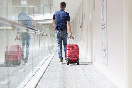 拖着行李旅行摄影照片_年轻男子拖着红色行李，在旅馆大楼里寻找自己的房间