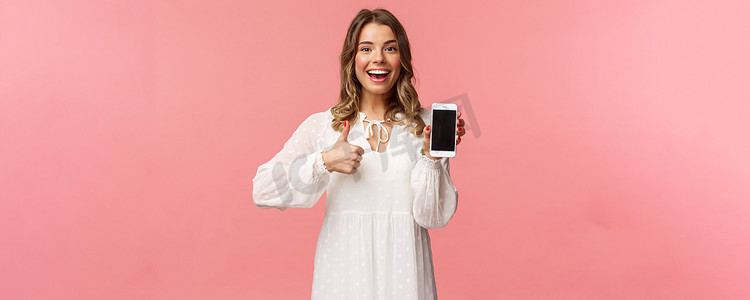 穿着白裙的乐观漂亮金发女性的肖像，推荐下载应用程序或订阅，使用在线购物应用程序，竖起大拇指，展示手机屏幕