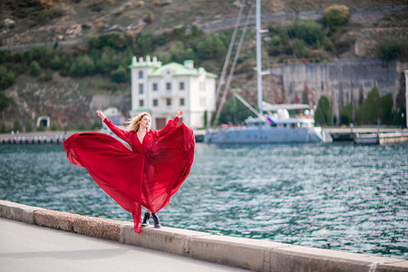 蓝色的裙子摄影照片_身着红裙的女子，长着丝绸长翅膀的时装模特，身着飘逸的裙子，在堤岸上飞舞着布料。