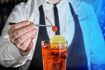一位专业酒保在夜总会柜台用玻璃酒吧镊子将樱桃放入红色冷冻酒精鸡尾酒中