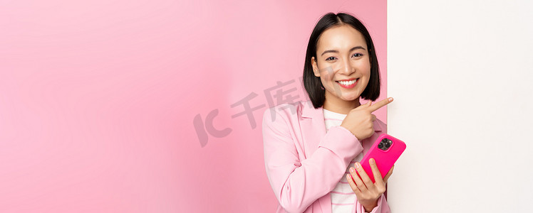 身穿西装、微笑的亚洲企业女性手持智能手机、指着董事会、在空荡荡的白墙上显示图表或信息标志、站在粉红色背景上的图片