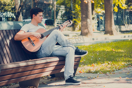 不的变调摄影照片_年轻人坐在公园的长椅上，用变调夹弹奏原声吉他。