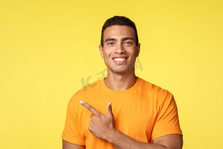 促销黄色背景摄影照片_特写年轻男性微笑，穿着 T 恤的快乐男人，指着左边，骄傲地打手势和咧嘴笑，展示促销或最佳选择，提供建议，推荐商店，站在黄色背景