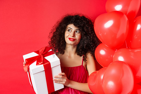 化妆和晚礼服的华丽女人，收到惊喜礼物和微笑，站在情人节浪漫气球附近的红色背景上