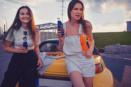 穿着休闲装的女孩正靠在黄色汽车的后备箱上，一边笑着享受玻璃瓶中的汽水，一边放着比萨饼。
