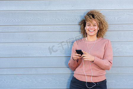 快乐的非洲裔美国女性站在灰色背景旁，用耳机和手机听音乐