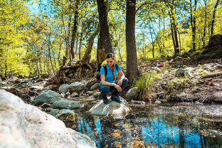 徒步旅行的女人坐在穿过树林的小溪中间的一块石头上